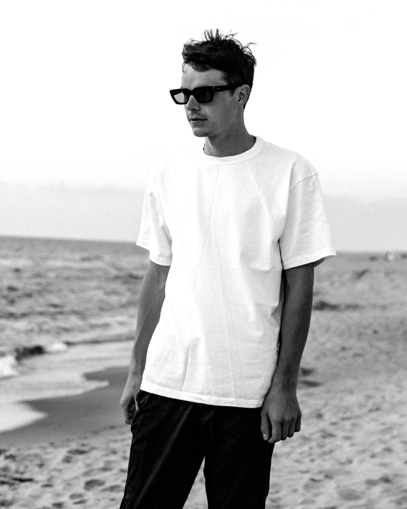 A portrait picture of Paulius Kairevicius standing next to the seaside wearing sunglasses, white t-shirt and black pants